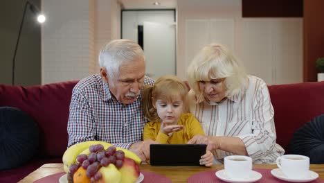 Abuelo-Y-Abuela-Sentados-En-La-Sala-De-Estar-Y-Enseñando-A-Su-Nieta-Pequeña-Usando-Una-Tableta