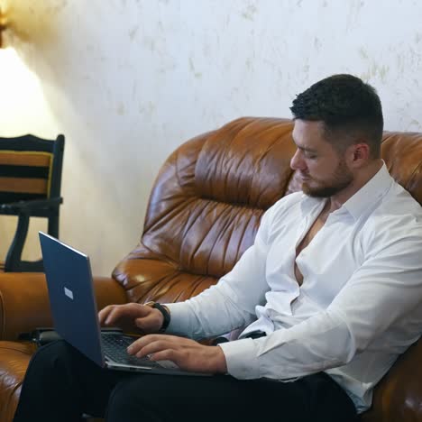 young man with a laptop sitting on sofa. elegant businessman working on a laptop on luxury couch in a hotel room. freelance, distance working