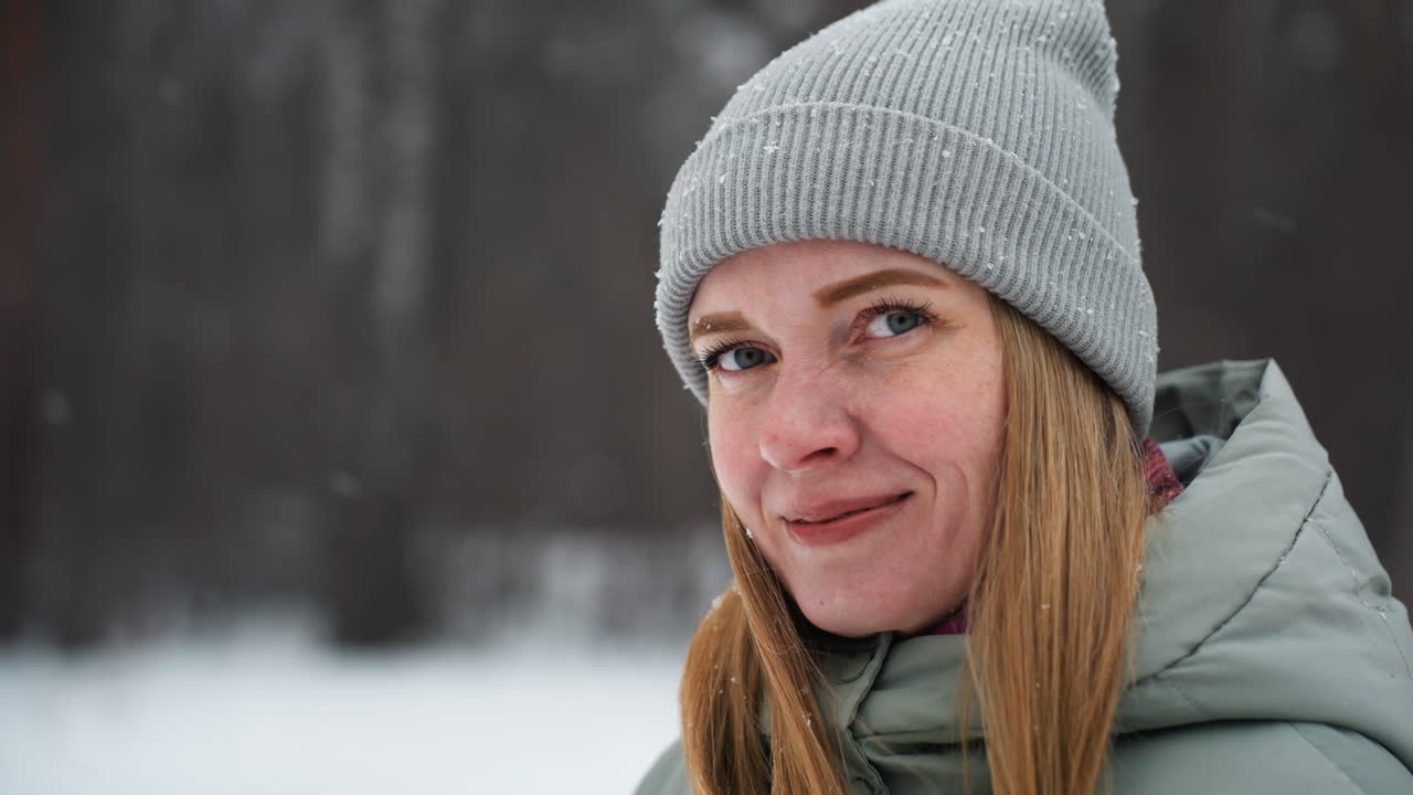 Primo piano di una donna con un berretto di lana grigio e una giacca invernale leggera che sorride dolcemente mentre è in piedi all'aperto in una foresta innevata, con fiocchi di neve che le cadono intorno al viso e alberi sfocati in uno sfondo stagionale freddo.