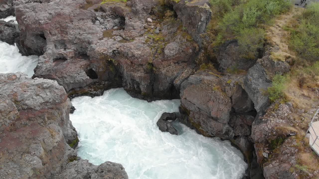 Flying over the flowing waterfall Barnafossar that's hitting the rocks and a tourist couple taking a selfie on the plateau with the waterfall in the background