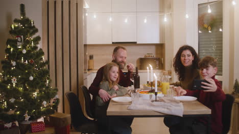 Der-Blonde-Junge,-Der-Mit-Seiner-Schwester-Und-Seinen-Eltern-Am-Weihnachtstisch-Sitzt,-Verwendet-Ein-Smartphone,-Um-Ein-Familien-Selfie-Zu-Machen