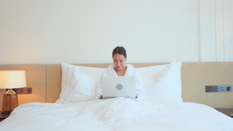 while snuggled up in a cozy bathrobe and kingsize bed, a young woman typing on her laptop proves she can work from anywhere and be successful