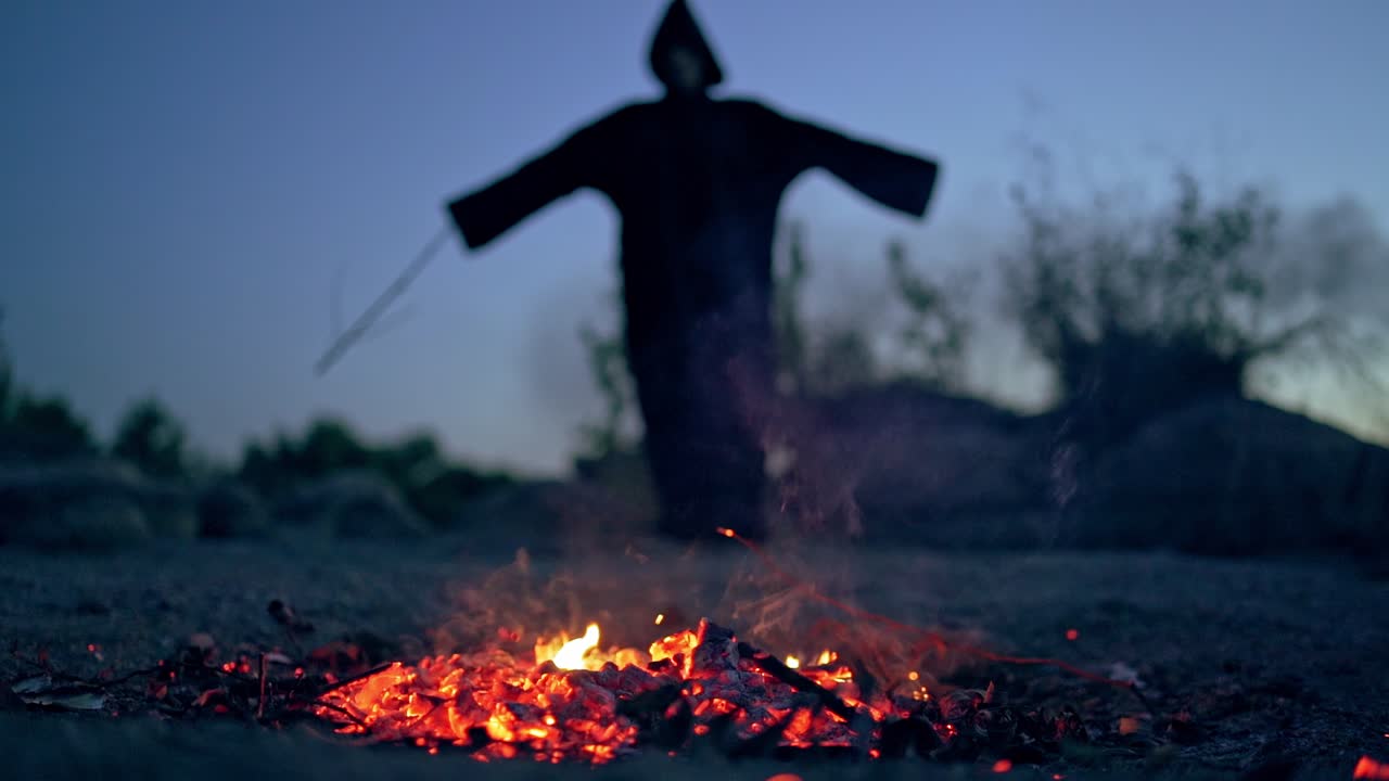 Embers of firewood and black scary silhouette. Mysterious figure in dark costume withwooden stick standing behind the smoldering logs of fire.