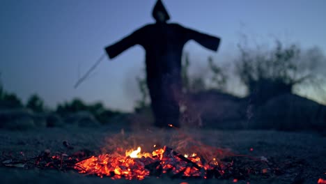 embers of firewood and black scary silhouette. mysterious figure in dark costume withwooden stick standing behind the smoldering logs of fire.