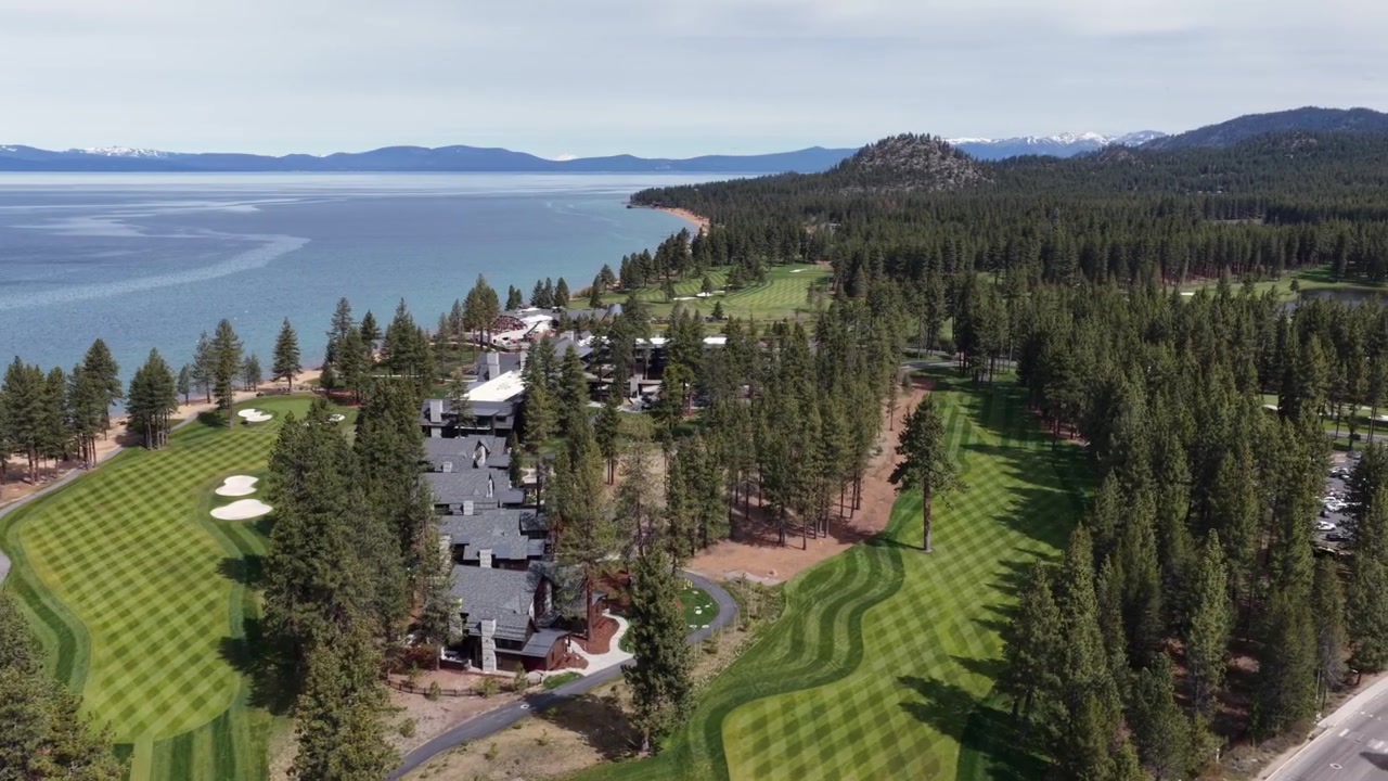 Golf course fairways and sand traps nestle among dense pine forest along turquoise Lake Tahoe shoreline with snow-dusted Sierra Nevada peaks beyond, California, USA. Aerial drone view.