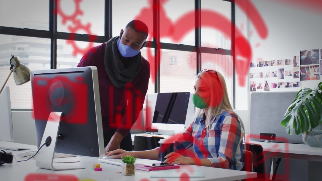 Animation of media icons over diverse business people wearing face masks and using computer