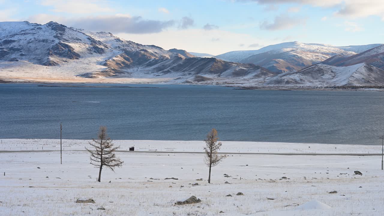 Remote Mongolia's seasonal change: A vast, blue lake is set against snow-covered mountains. A snowy steppe with sparse larches and a road highlights the stark transition from autumn to winter
