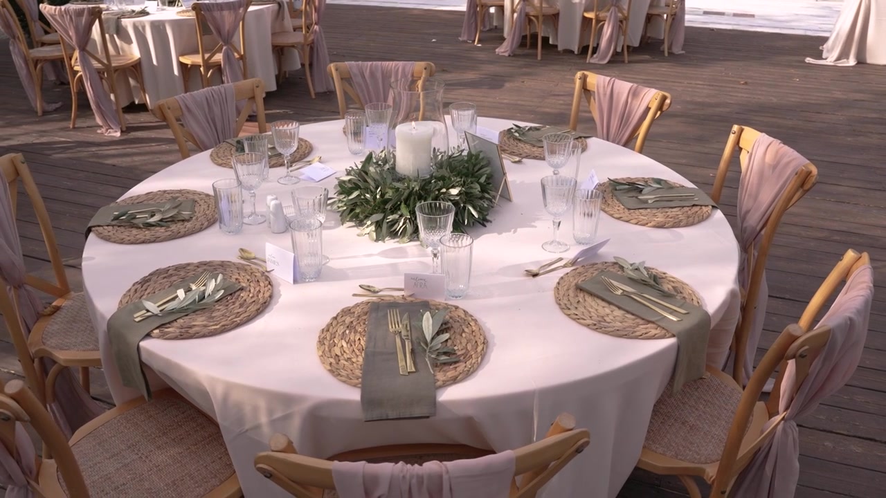 The table is set for a wedding reception. It features woven placemats, glasses, and a centerpiece with greenery. Chairs are around the table in an outdoor area.