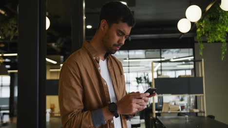 hombre de negocios biracial de pie usando un teléfono inteligente en una oficina moderna