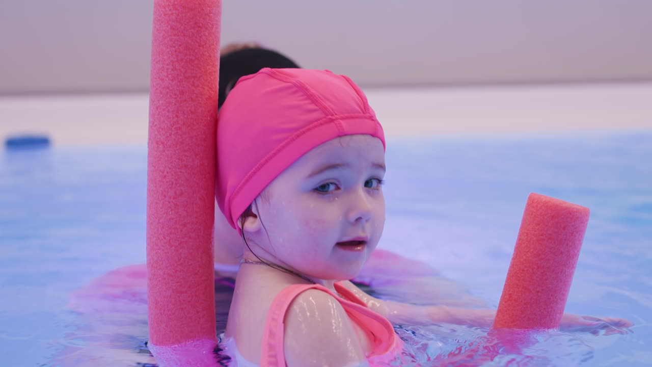 Close view of an infant resting on foam guard while floating in water, turning head slightly left with calm relaxed expression, delicate features highlighted under soft pool lighting