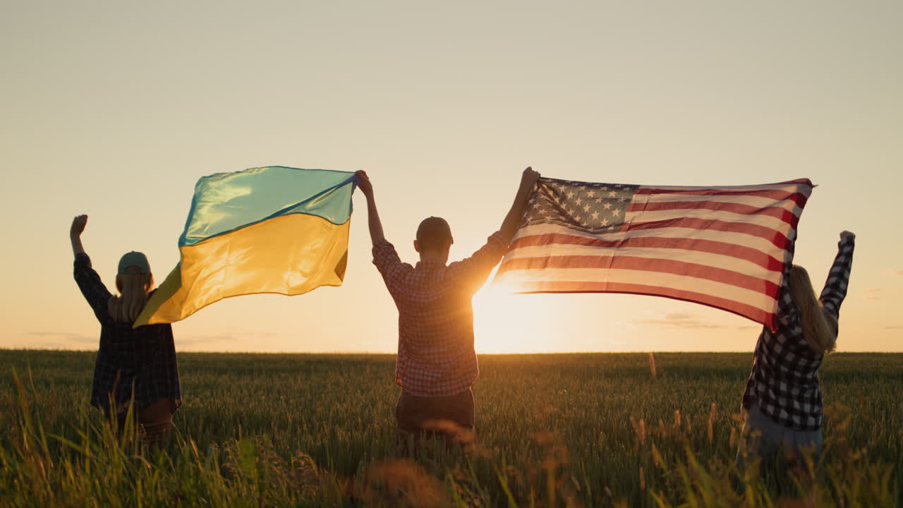 A group of friends raises the flag of the United States and the flag of Ukraine over their heads. Standing against the backdrop of a field of wheat where the sun sets
