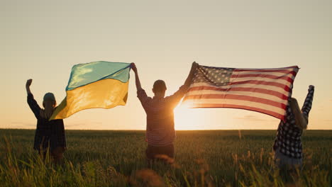 un grupo de amigos levanta la bandera de los estados unidos y la bandera de ucrania sobre sus cabezas. de pie contra el telón de fondo de un campo de trigo donde el sol se pone