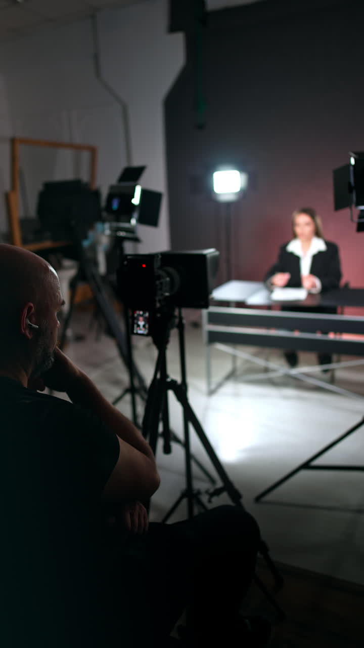 Rear view of a cameraman sitting at camera. Videographer taking video of a woman sitting at desk. Vertical video.