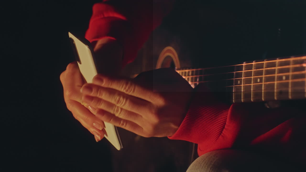 Closeup of woman tuning guitar with device. Detailed image of woman tuning guitar with phone in firelight. Woman finetunes guitar strings with phone under campfire glow while focusing closely