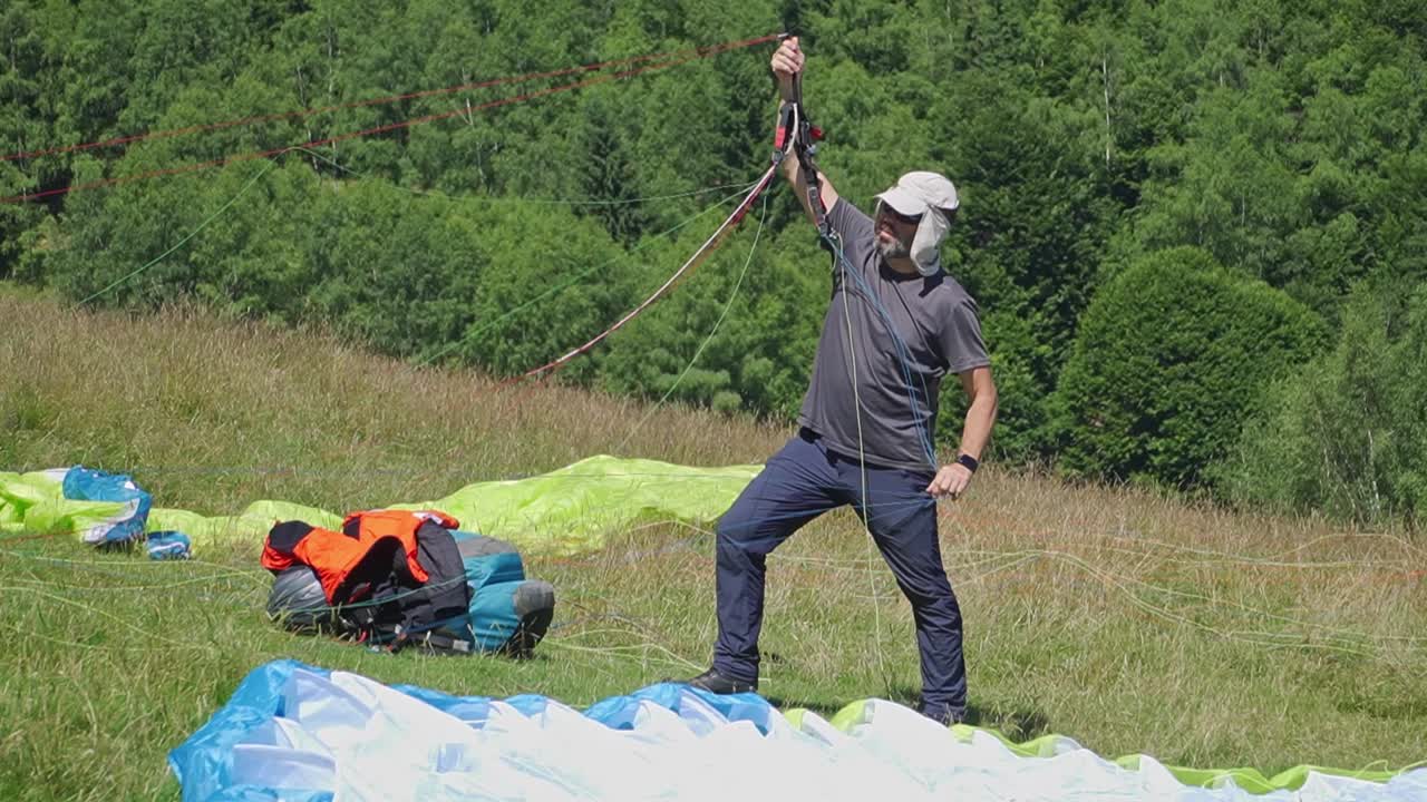 A man focuses on getting his parachute ready, carefully handling the wires before taking off into the sky. Another parachute in the background. Extreme adventure