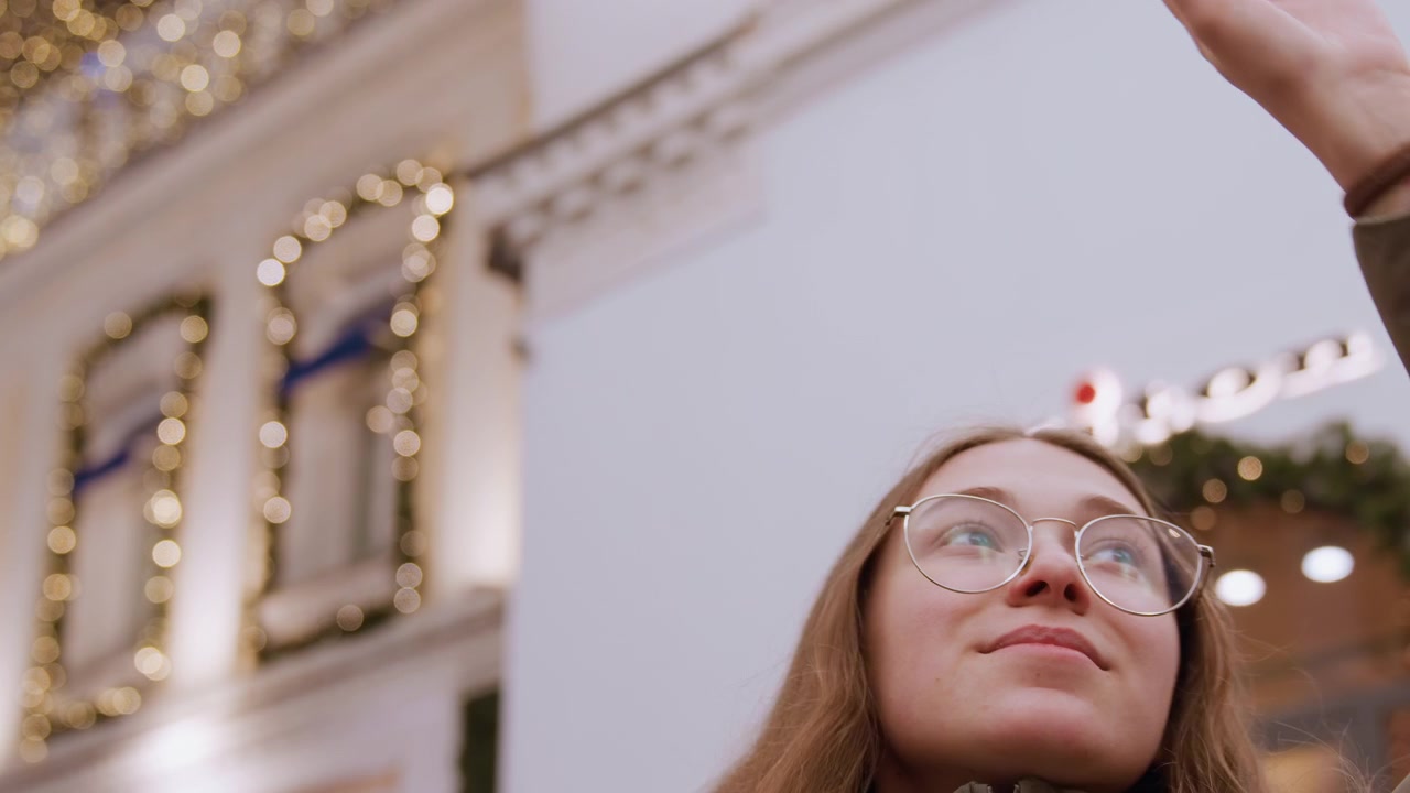 Giovane donna che si protende sotto un baldacchino di luci dorate e ghirlande, con una ghirlanda da vetrina e una facciata ornata sullo sfondo. Una acquirente natalizia che indossa un piumino alza la mano, con espressione deliziata e occhiali.