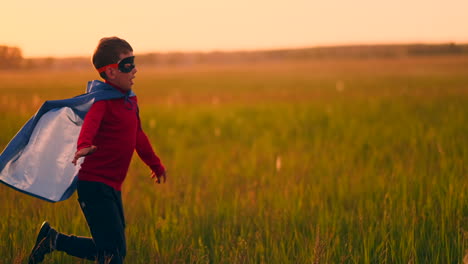 un niño con un traje y una máscara de superhéroe corre a través del campo al atardecer en la hierba soñando e imaginándose a sí mismo un héroe y un ganador. familia divertida y feliz