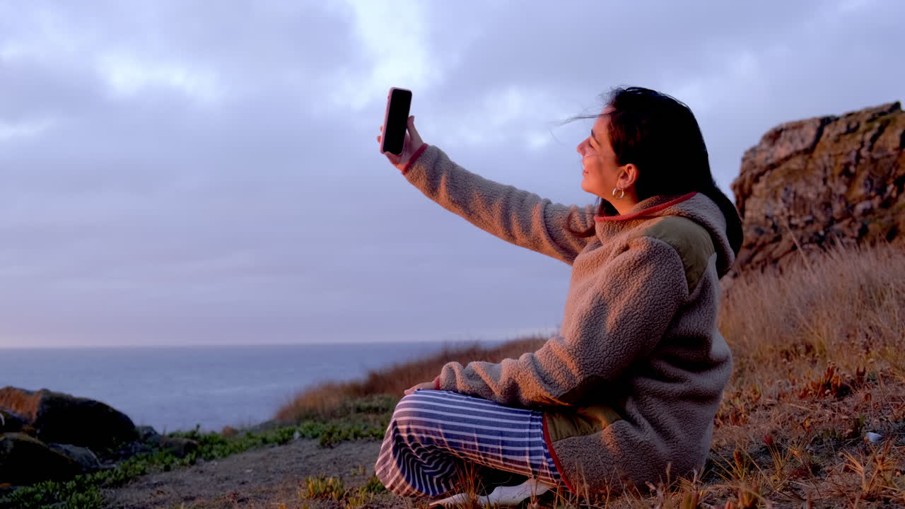 mujer mirando la cámara de su teléfono celular, saludando y sonriendo, puesta de sol en la playa, océano pacífico chile buchupureo