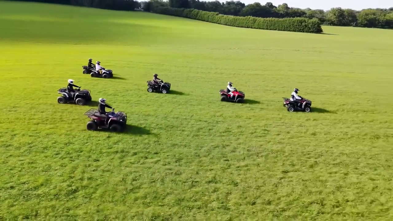 Diversi piloti di quad attraversano un ampio campo erboso in aperta campagna sotto un sole intenso, visti da un punto di vista aereo elevato con lunghe ombre e alberi in lontananza.