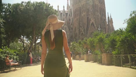 stunning video of a young caucasian female in a green dress with a knitted hat, walking to incredible landmark building - sagrada familia, barcelona