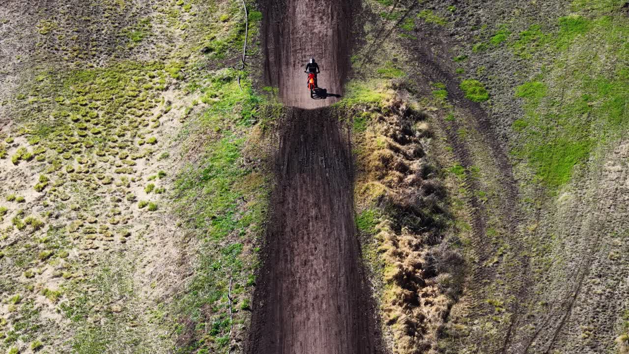 Motocross rider speeds along rural dirt track, aerial perspective, bright daylight, dynamic movement