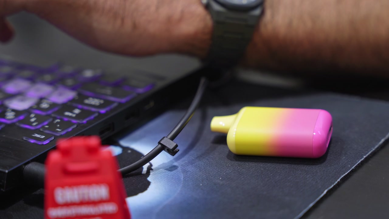 Primo piano di una sigaretta elettronica usa e getta rosa e gialla su una scrivania accanto a un laptop. Una persona lavora sullo sfondo vicino a un segnale di avvertimento sfocato in un moderno ambiente d'ufficio.