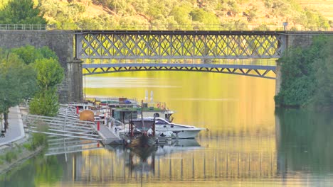 Barcos-Atracados-Frente-A-Un-Puente-De-Hierro-Que-Cruza-El-Río-Duero-En-Pinhao,-Portugal.