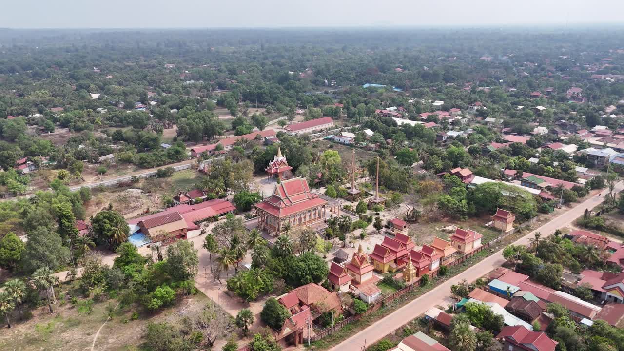 Traditional Khmer architecture with its ornate, tiered roofs and golden accents. Several smaller structures and perhaps residential buildings with red tile roofs are clustered around the temple.