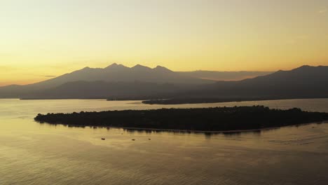 Misty-yellow-sky-after-sunset-over-mountains-horizon-reflecting-sunlight-on-calm-sea-surface-around-silhouette-of-tropical-island,-Bali