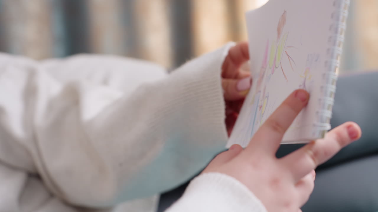 Hands holding sketchbook with colorful child drawing of house, tree, sun and people, finger pointing at detail of artwork, cozy home atmosphere with soft fabric background and natural warm lighting