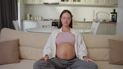 portrait of a happy brunette pregnant woman who sits on the sofa and meditates while she is expecting a baby at home