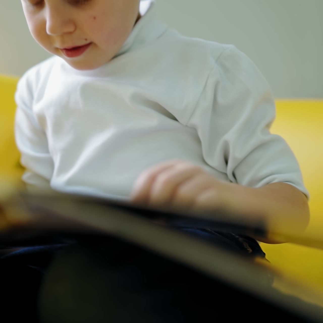 Little boy with a big book. Preschool boy sitting alone on the couch and reading book turning pages at home. Close-up.