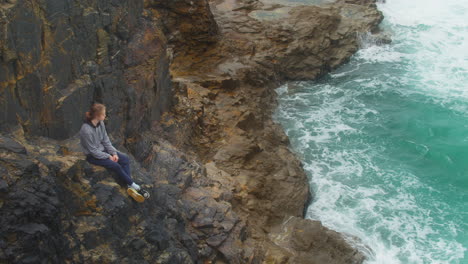 Sad-Young-Man-Is-Sitting-On-Cliff-Edge-Looking-At-The-Ocean-In-Cornwall,-England