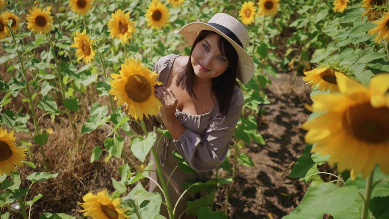 Donna con un cappello a tesa larga interagisce con i girasoli in un campo, tenendo e annusando i fiori, circondata da foglie verdi e fiori gialli. La scena è caratterizzata da una luce solare intensa.