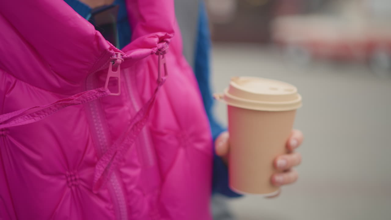 Primo piano di una donna che tiene in mano un bicchiere di caffè da asporto, giubbotto imbottito rosa con dettagli a cerniera, una sola mano che stringe un bicchiere di carta caldo, sfondo sfocato di strada cittadina, atmosfera da pendolarismo con clima fresco, abbigliamento casual