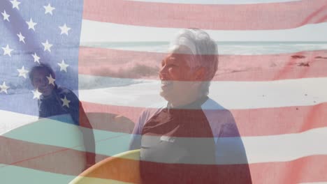 animation of flag of united states of america over senior biracial couple with surfboards on beach