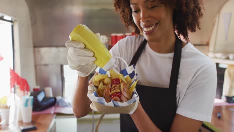 Mujer-Afroamericana-Con-Delantal-Poniendo-Mostaza-Sobre-El-Hot-Dog-En-Un-Camión-De-Comida