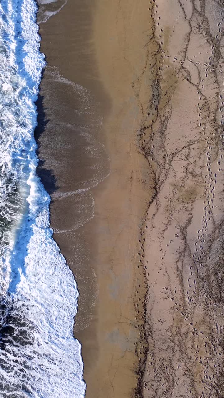 Inquadratura cinematografica verticale 9:16 da drone di onde marine che si infrangono sulla riva. Vista dall'alto ad angolo acuto di texture di spiaggia sabbiosa, schiuma dell'oceano e impronte. Perfetto per reel di viaggio sui social media