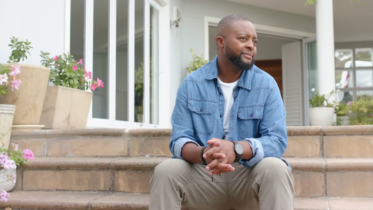 African American man sitting on steps, hands clasping, shifting gaze right, smiling near planters