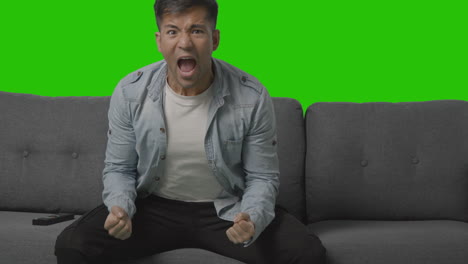 studio shot of young man celebrating sitting on sofa watching sport on tv against green screen
