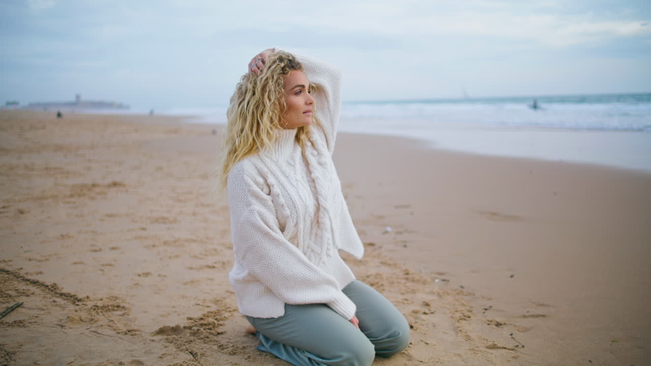 Woman resting autumn beach on cloudy day. Beautiful curly tourist looking camera