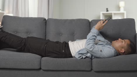 young man lying on sofa at home scrolling through mobile phone