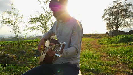 Músico-Masculino-Asiático-Tocando-La-Guitarra-Mientras-Se-Sienta-Con-El-Sol-Brillando-Y-Bengalas,-Vietnam