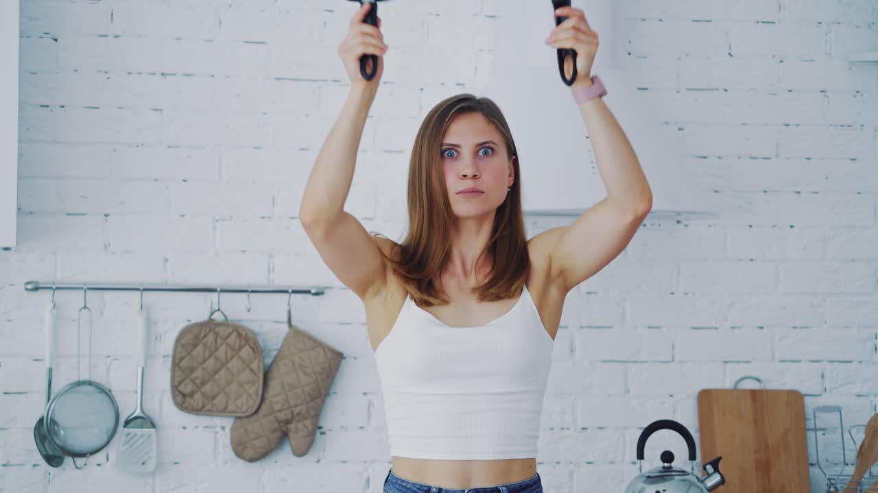 Woman with new frying pans in the kitchen. Beautiful female holding two empty black frying pans. Happy girl showing different emotions on her face in kitchen.