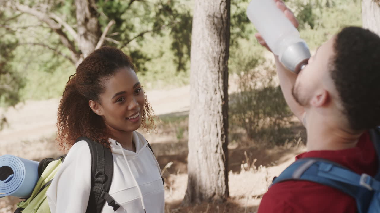 Happy african american couple wearing backpacks and drinking water in forest, slow motion