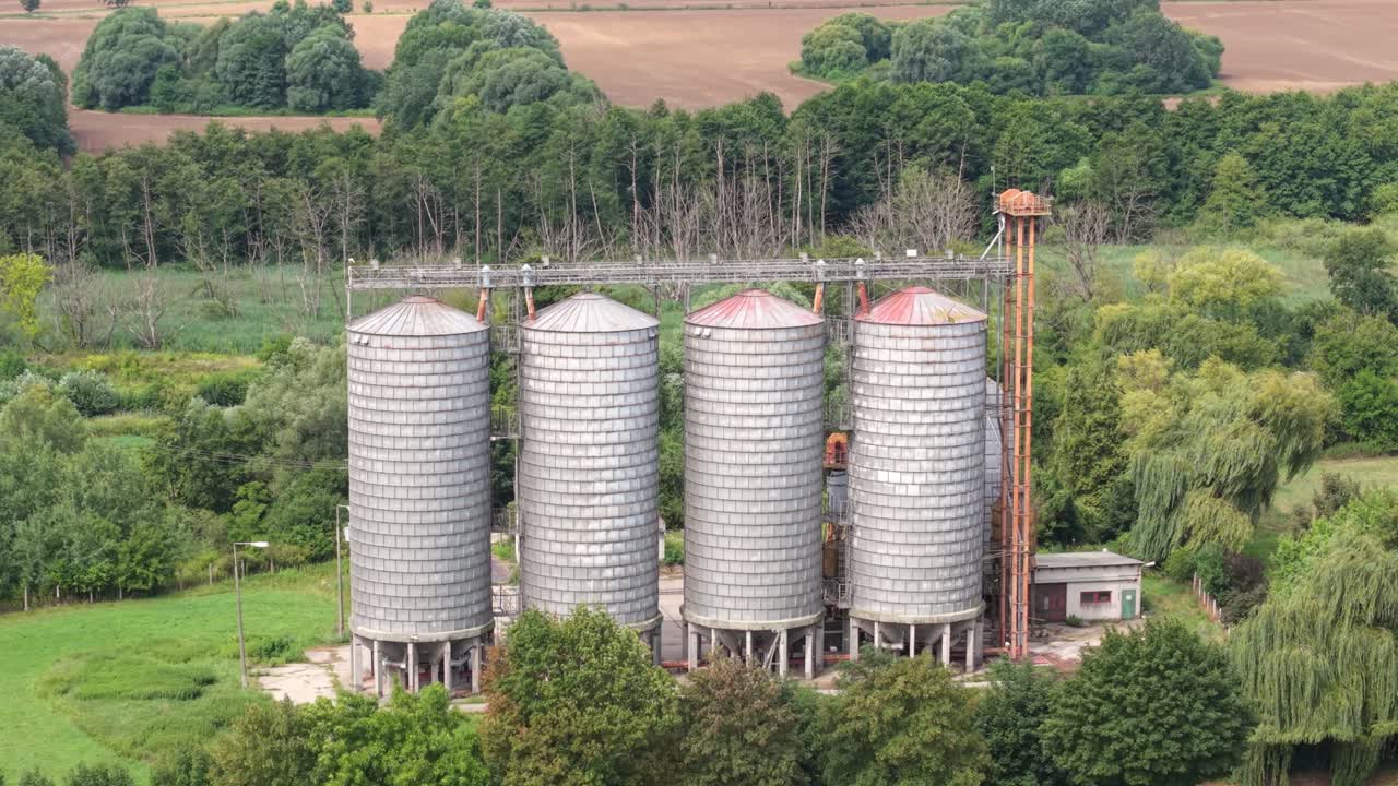 A set of large cylindrical grain silos stands surrounded by lush greenery and forest on the edge of an agricultural field. The structures are used for storing harvested grain
