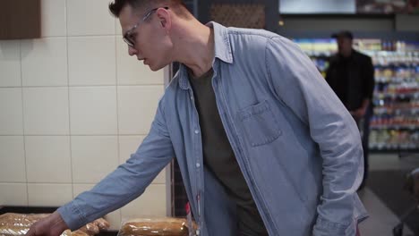 Close-up-of-a-caucasian-stylish-man-choosing-the-best-bread-in-a-supermarket-from-a-bakery-counter.-Chose-a-small-french-baguette-for-a-dinner.-Side-view