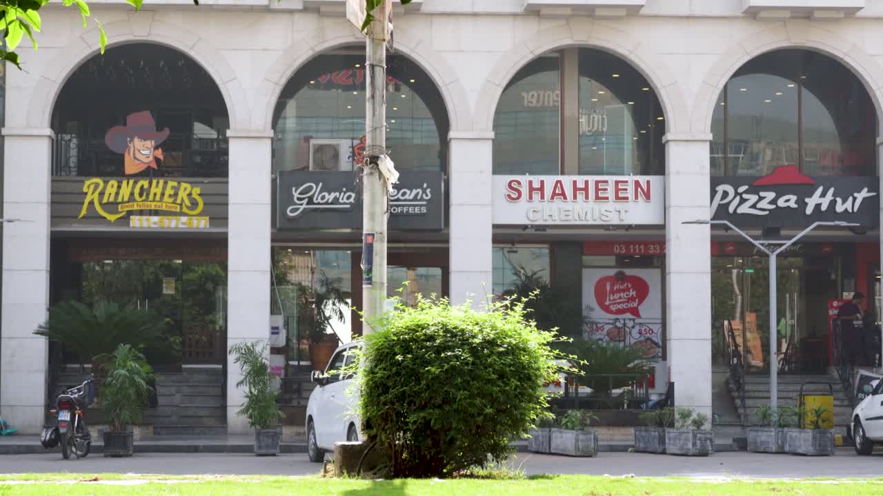 A view of a bustling street, with towering commercial buildings, restaurants, and a chemist store standing proudly amidst the hustle and bustle of passing cars.