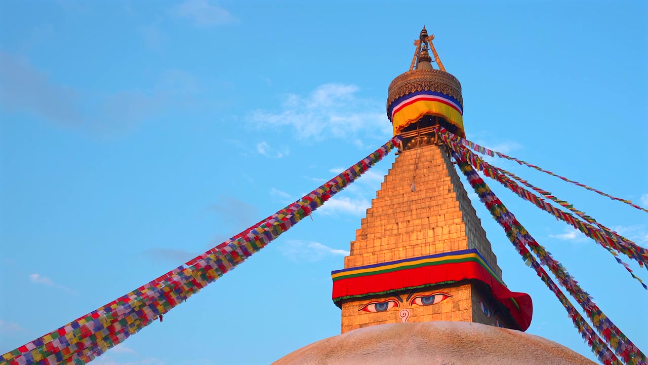 Vista dal basso dell'iconica guglia dello Stupa di Boudhanath decorata con bandiere di preghiera buddiste colorate che sventolano dolcemente al vento contro un cielo blu al tramonto a Kathmandu, Nepal.