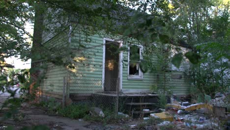 dilapidated wooden house with broken windows and no door and huge amounts of garbage in the yard among the green trees in detroit